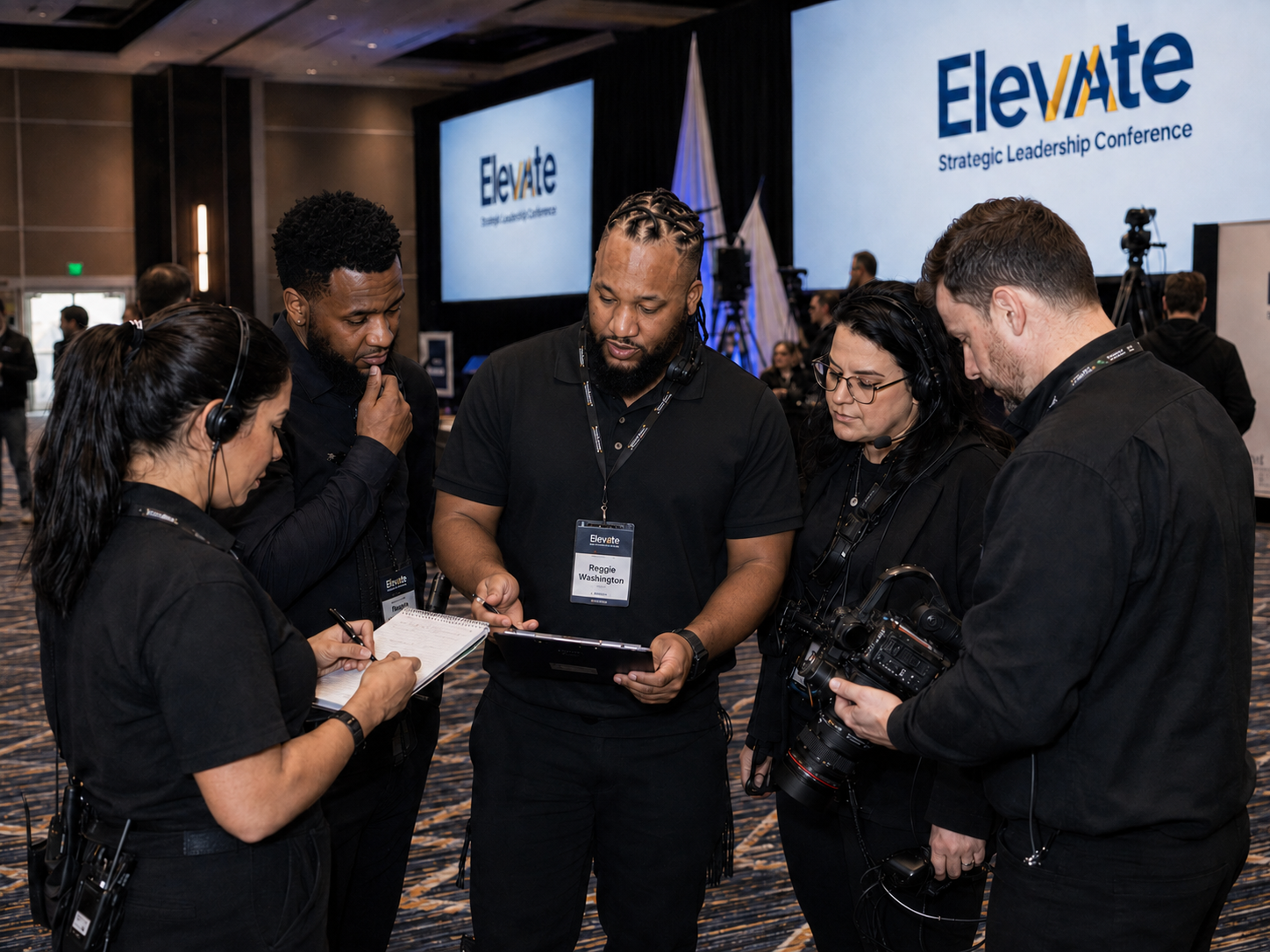 Reggie Washington briefing a five person crew on a tablet at a leadership conference, camera operators and headsets around him